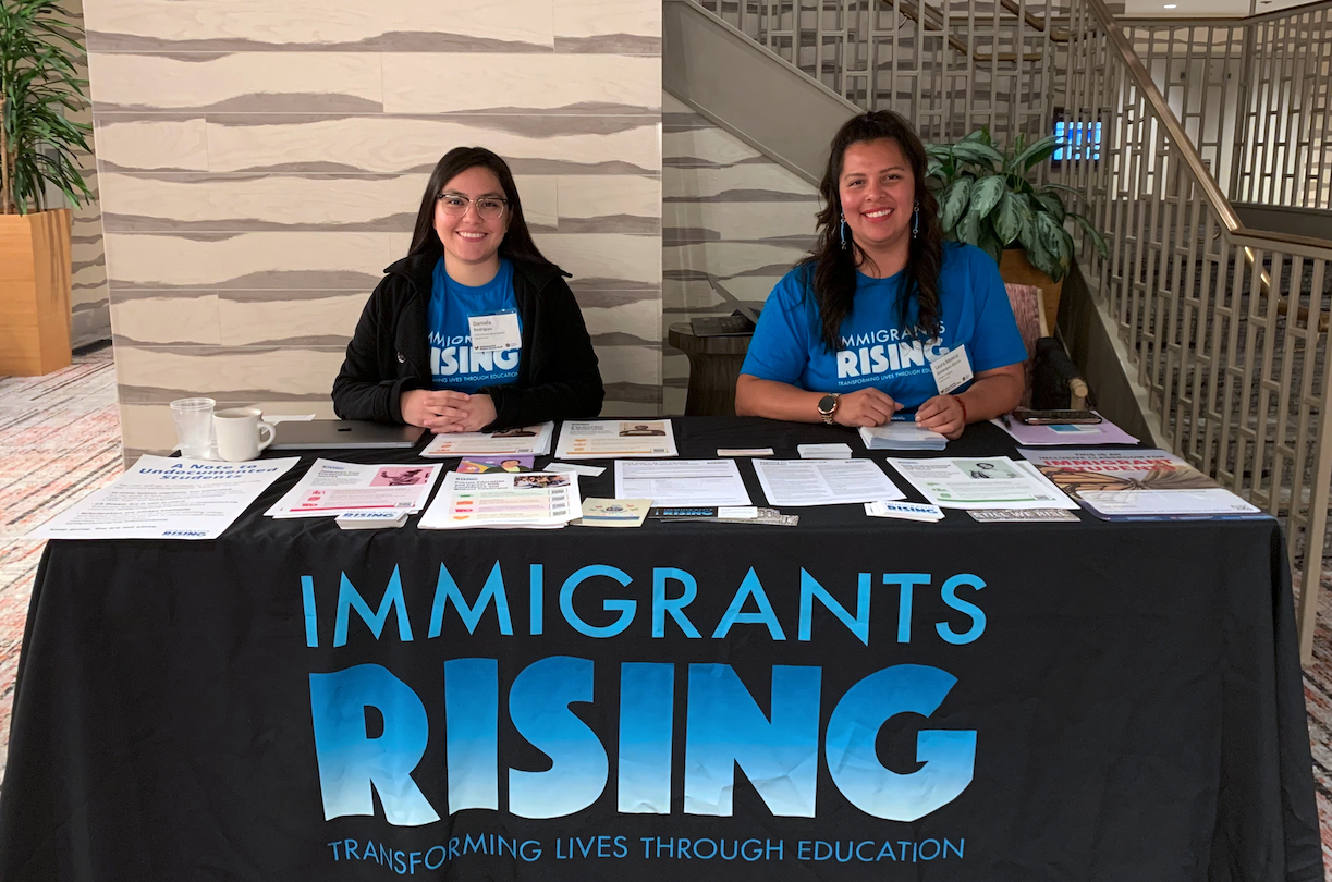 Immigrants Rising team members sit behind a resource table with flyers, resources, and swags.